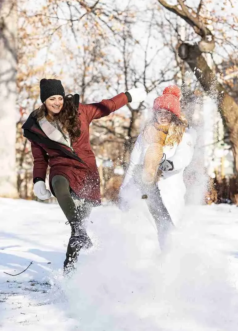 Två glada kvinnor i mössa och vinterjackor sparkar upp snö framför en skog.