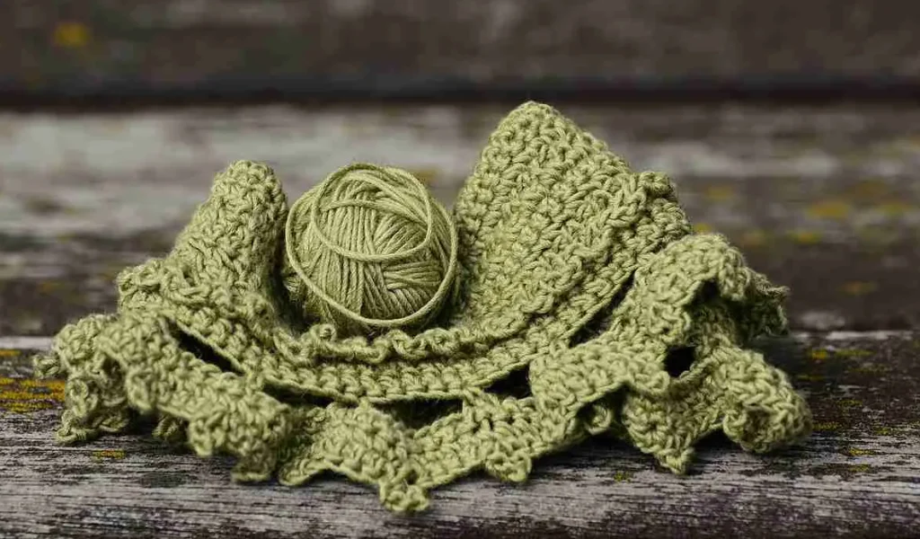 En virkad grön sjal ligger ihopvikt på en träbänk, med ett garnnystan i samma färg som ligger ovanpå.